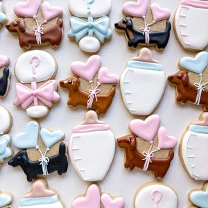 May include: Assortment of decorated sugar cookies, including dachshunds with heart-shaped balloons, baby bottles, and cookies with question marks and bows. The cookies are decorated with pink, blue, brown, black, and white icing, perfect for a baby shower.