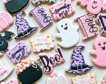 May include: A collection of Halloween-themed sugar cookies decorated with various designs, including ghosts, pumpkins, bats, witches' hats, and cauldrons. Some cookies feature the text "Boo!" and "Millie is a spooky one".