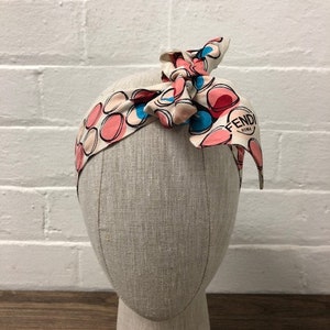 Op de afbeelding: Een wit hoofdband met een roze, blauwe en witte stippenpatroon. Het hoofdband heeft een strik bovenop en de tekst "FENDI ROMA" is aan de zijkant gedrukt.