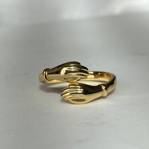 May include: A gold ring with two hands clasped together. The ring is designed to be worn on the finger.