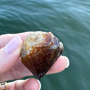 Peut inclure: Un cristal ambré facetté avec une base marron foncé. Le cristal est tenu dans une main, avec une bague en or visible. L'arrière-plan est une vue floue de l'eau.