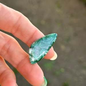 Peut inclure: Un cabochon d'agate vert et blanc en forme de larme. La pierre est translucide avec des tourbillons de vert et de blanc.