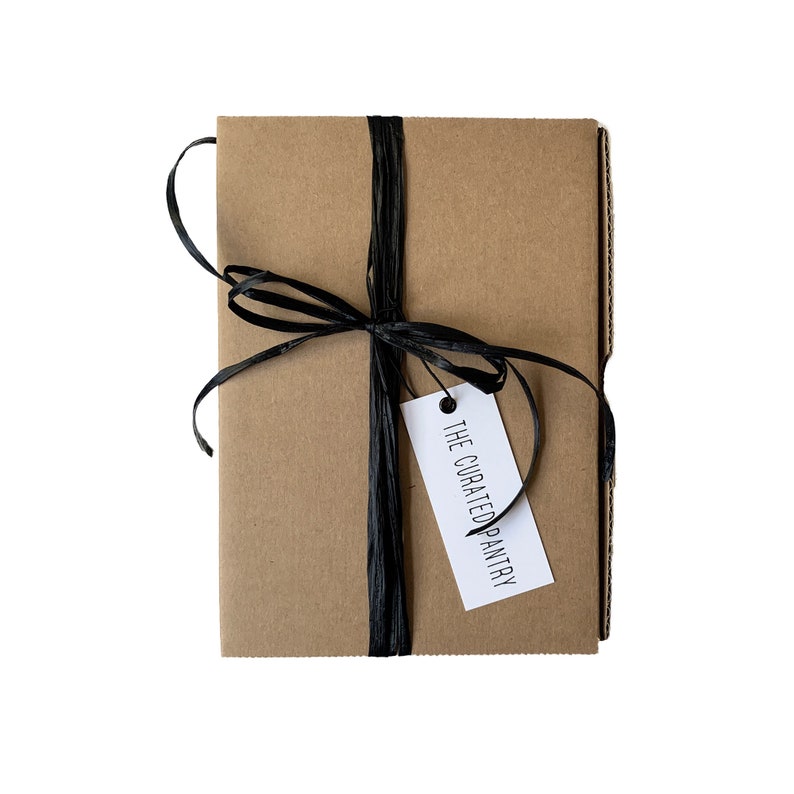 Puede incluir: Una caja de cart&oacute;n marr&oacute;n atada con una cinta negra y una etiqueta blanca que dice "THE CURATED PANTRY".