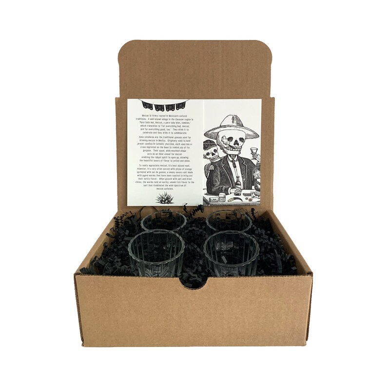 Puede incluir: Un juego de seis vasos de chupito de vidrio transparente en una caja de cart&oacute;n marr&oacute;n con papel triturado negro. La caja est&aacute; abierta y los vasos son visibles. La caja tiene una tarjeta dentro con una ilustraci&oacute;n en blanco y negro de tres esqueletos bebiendo y un texto sobre el tequila.