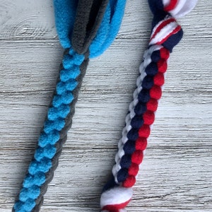 May include: Two dog toys made of fleece. One is blue and gray, with a braided handle and a fringed top. The other is red, white, and blue, with a braided handle and a fringed top. The toys are designed for dogs.