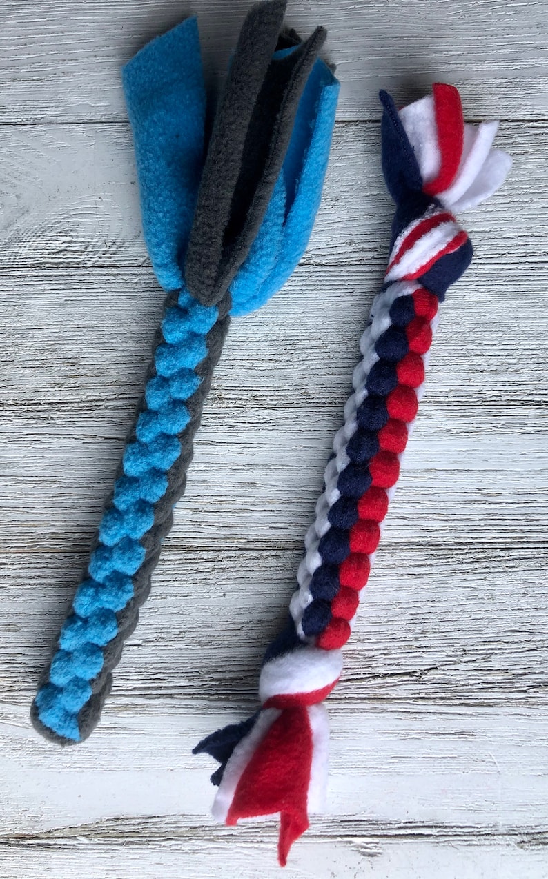 May include: Two dog toys made of fleece. One is blue and gray, with a braided handle and a fringed top. The other is red, white, and blue, with a braided handle and a fringed top. The toys are designed for dogs.