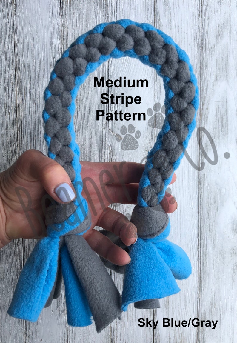 May include: Sky blue and gray dog toy with a braided rope design. The toy features a medium stripe pattern and fleece tassels at both ends. The text "Medium Stripe Pattern" and "Sky Blue/Gray" are visible.