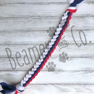May include: Braided dog toy in red, white, and blue. The toy is made of soft fabric and features a knotted end and a tassel of fabric strips. The text "Beamer and Co." is visible on the background.