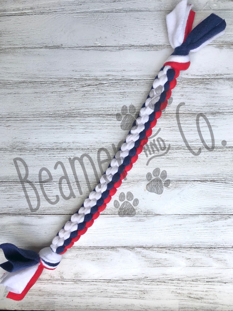 May include: Braided dog toy in red, white, and blue. The toy is made of soft fabric and features a knotted end and a tassel of fabric strips. The text "Beamer and Co." is visible on the background.