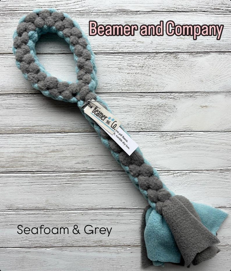 May include: A braided dog toy in seafoam and grey. The toy has a loop at the top and a long, braided body that ends in two flaps of fleece. The text "Beamer and Company" and "Seafoam & Grey" are visible.