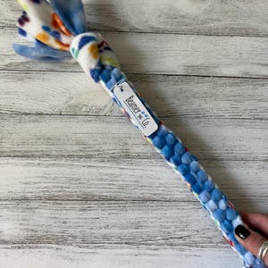 May include: A blue and white dog toy with a knotted end and a braided body. The knotted end features colorful spots, while the body is a woven pattern of blue fabric. A tag reads "Beamer & Co."