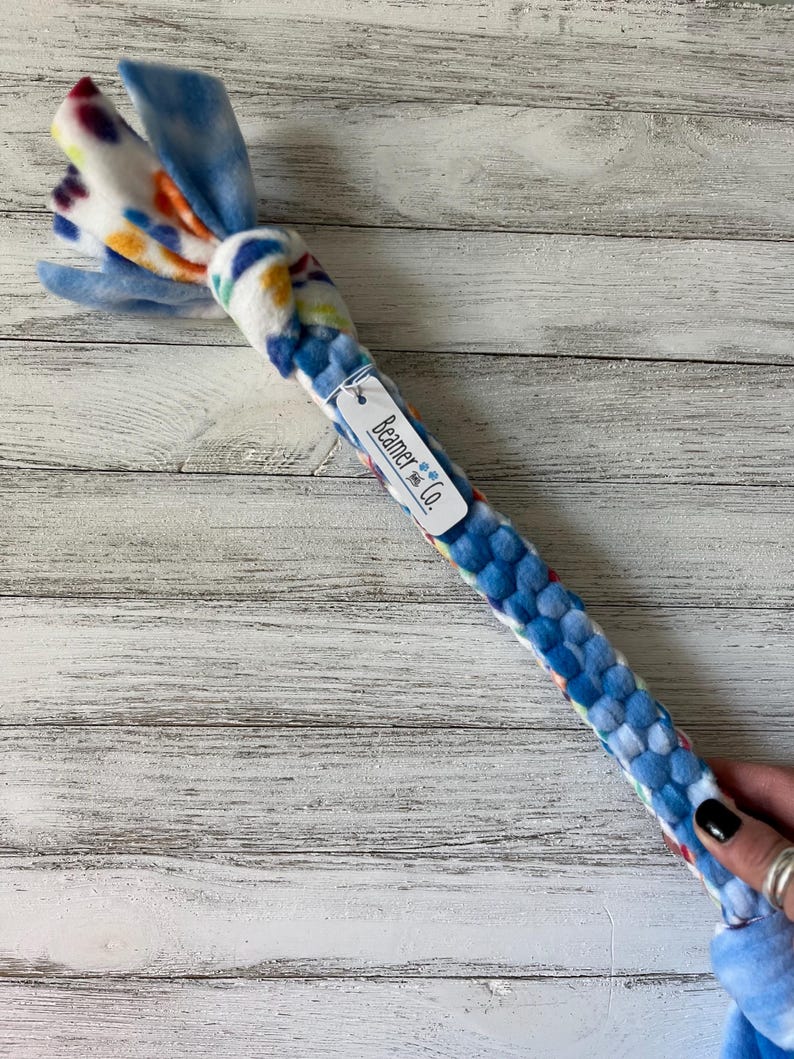 May include: A blue and white dog toy with a knotted end and a braided body. The knotted end features colorful spots, while the body is a woven pattern of blue fabric. A tag reads "Beamer & Co."