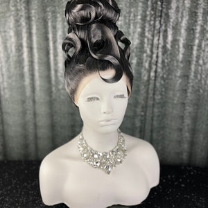 May include: A black wig with a high, elaborate updo styled with large, dramatic curls. The wig is on a mannequin head with a white bust and a silver rhinestone necklace.