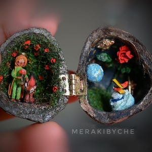 May include: A miniature scene inside a hinged walnut shell, featuring a small figurine of The Little Prince with a fox, a red rose, and a blue planet. The scene is surrounded by green moss and red flowers.