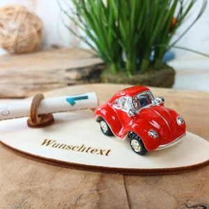 Könnte beinhalten: Ein kleines rotes Spielzeugauto mit silbernen Akzenten steht auf einer Holzplakette. Auf der Plakette steht der deutsche Text "Wunschtext". Eine gerollte Schriftrolle mit einem blauen Pfeil wird von einem Holzhalter gehalten.