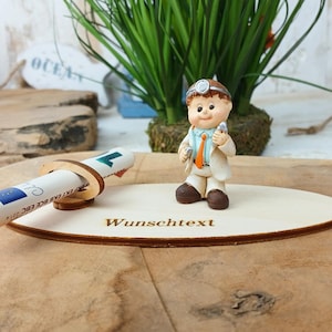 Könnte beinhalten: Figur eines Arztes im weißen Kittel, der medizinische Instrumente hält, steht auf einem ovalen Holzsockel. Eine aufgerollte Banknote wird in einem Holzhalter gehalten. Der Text "Wunschtext" ist auf das Holz graviert.