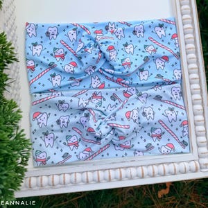 May include: Two light blue headbands with a repeating pattern of cartoon teeth wearing Santa hats, scarves, and holding toothbrushes and candy canes. The headbands are twisted at the top.