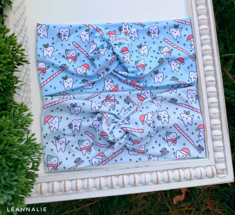 May include: Two light blue headbands with a repeating pattern of cartoon teeth wearing Santa hats, scarves, and holding toothbrushes and candy canes. The headbands are twisted at the top.