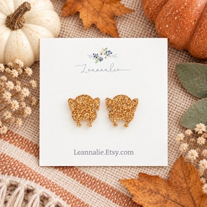 May include: A pair of gold glitter turkey-shaped stud earrings attached to a white display card. The card features a small floral graphic, the brand name Leannalie in cursive script, and the website address Leannalie.Etsy.com printed in black.