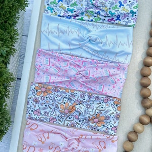 K&ouml;nnte beinhalten: F&uuml;nf bunte Stirnb&auml;nder mit verschiedenen Mustern. Das oberste Stirnband hat einen wei&szlig;en Hintergrund mit rosa, blauen, gr&uuml;nen und gelben B&auml;ndern mit den Worten "HOPE" und "CURE". Das zweite Stirnband hat einen hellblauen Hintergrund mit einem rosa EKG-Muster. Das dritte Stirnband hat einen rosa Hintergrund mit einem wei&szlig;en Muster aus Stethoskopen und Herzen. Das vierte Stirnband hat einen wei&szlig;en Hintergrund mit einem rosa, orangefarbenen und gelben Muster aus Blumen, Stethoskopen und Spritzen. Das f&uuml;nfte Stirnband hat einen rosa Hintergrund mit einem wei&szlig;en Muster aus Stethoskopen und Blumen.