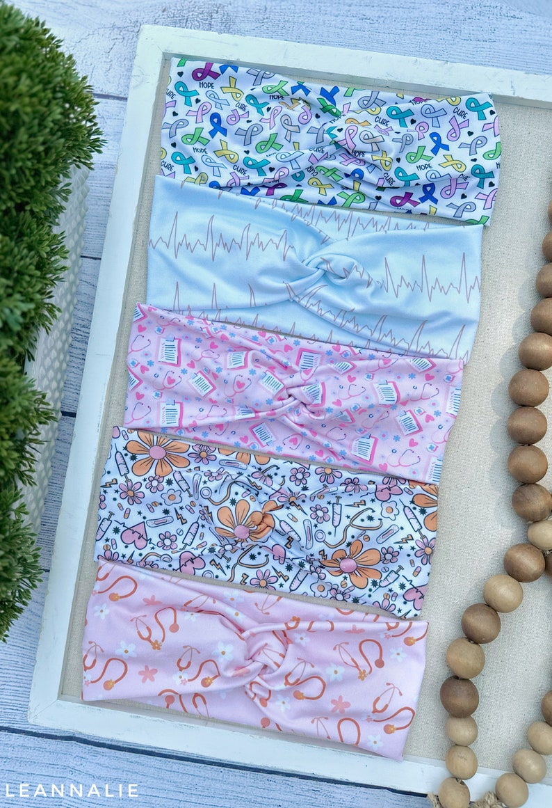 K&ouml;nnte beinhalten: F&uuml;nf bunte Stirnb&auml;nder mit verschiedenen Mustern. Das oberste Stirnband hat einen wei&szlig;en Hintergrund mit rosa, blauen, gr&uuml;nen und gelben B&auml;ndern mit den Worten "HOPE" und "CURE". Das zweite Stirnband hat einen hellblauen Hintergrund mit einem rosa EKG-Muster. Das dritte Stirnband hat einen rosa Hintergrund mit einem wei&szlig;en Muster aus Stethoskopen und Herzen. Das vierte Stirnband hat einen wei&szlig;en Hintergrund mit einem rosa, orangefarbenen und gelben Muster aus Blumen, Stethoskopen und Spritzen. Das f&uuml;nfte Stirnband hat einen rosa Hintergrund mit einem wei&szlig;en Muster aus Stethoskopen und Blumen.