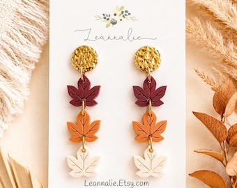 Pode incluir: Brincos pendentes com um pino circular dourado texturizado conectado a três pingentes de folha de bordo empilhados nas cores bordô, laranja e creme. O cartão de exibição branco apresenta o texto Leannalie e Leannalie.Etsy.com com um logotipo floral.