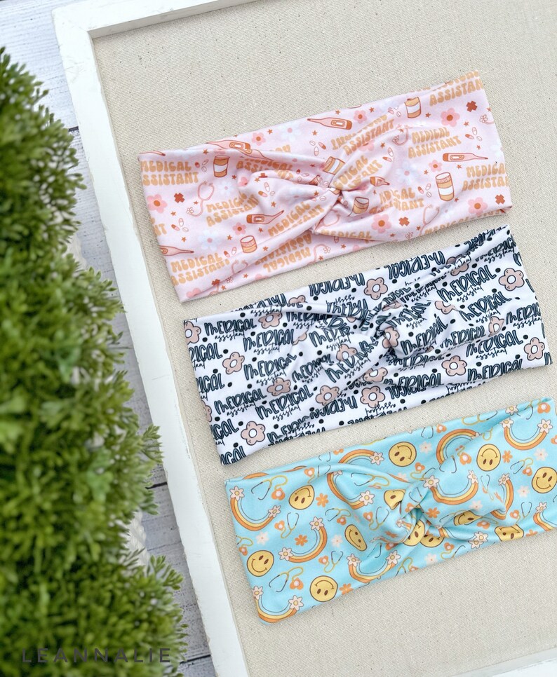 May include: Three colorful headbands with different patterns. The first headband is pink with a white pattern of medical symbols and the text "Medical Assistant". The second headband is white with a black pattern of flowers and the text "Medical". The third headband is light blue with a white pattern of rainbows, smiley faces, and stethoscopes.