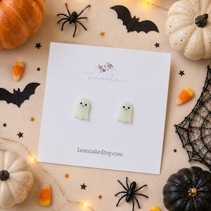 Puede incluir: Un par de pendientes con forma de fantasma con caras sonrientes en una tarjeta blanca. La tarjeta tiene el texto "Leannalie.Etsy.com" y está rodeada de decoraciones de Halloween, incluyendo calabazas, arañas y caramelos de maíz.