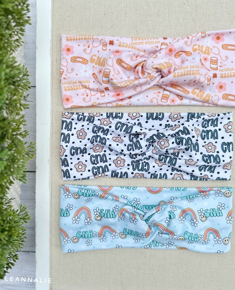 May include: Three colorful headbands with different patterns. The top headband is pink with a white background and features a repeating pattern of stethoscopes, pills, bandages, and the text "CNA Certified Nursing Assistant". The middle headband is white with a black background and features a repeating pattern of the text "CNA". The bottom headband is light blue with a white background and features a repeating pattern of rainbows, flowers, and the text "CNA".