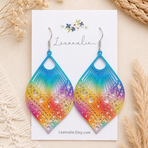 May include: A pair of dangle earrings featuring a diamond-shaped, intricate filigree design with a vibrant rainbow gradient of blue, yellow, orange, and pink. The earrings are displayed on a white card labeled Leannalie and Leannalie.Etsy.com.