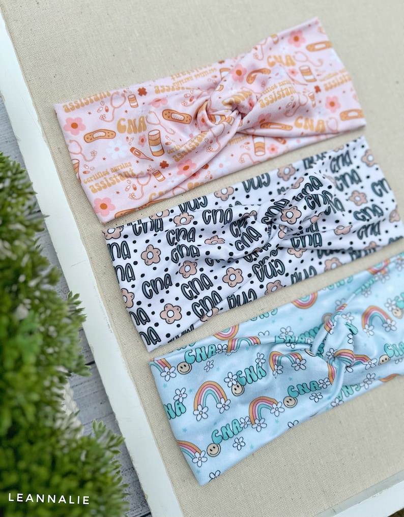 May include: Three colorful headbands with different patterns. The first headband is pink with a white background and features a repeating pattern of medical symbols, including a stethoscope, bandages, pills, and the text "Certified Nursing Assistant" and "CNA". The second headband is white with a black background and features a repeating pattern of black dots, white flowers, and the text "CNA". The third headband is light blue with a white background and features a repeating pattern of rainbows, white flowers, and the text "CNA".