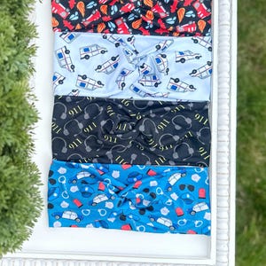 May include: Five fabric headbands with a twisted knot design. The headbands feature various patterns: red fire trucks on black, ambulances on light blue, 911 and headphones on black, and police cars and handcuffs on blue. The headbands are displayed on a white tray.
