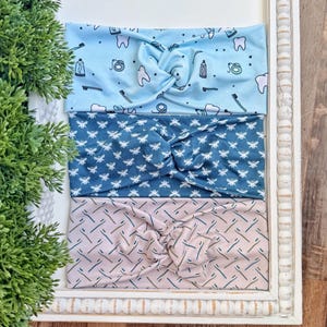 Könnte beinhalten: Drei Stoff-Haarbänder mit einem verdrehten Knoten-Design. Das obere Haarband ist hellblau mit zahnmedizinischen Grafiken. Das mittlere Haarband ist dunkelblau mit einem weißen Bienenmuster. Das untere Haarband ist rosa mit einem türkisfarbenen diagonalen Linienmuster.