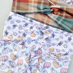 May include: Three fabric headbands with different patterns. The top headband is a plaid pattern in shades of brown, green, and orange. The middle headband is a white fabric with a pink and orange pumpkin and flower pattern. The bottom headband is a white fabric with a pink and orange pumpkin and flower pattern.