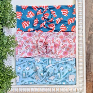 Könnte beinhalten: Drei Stoff-Stirnbänder mit einem verdrehten Knoten-Design. Eines ist türkis mit roten und weißen Zähnen, ein anderes ist rosa mit Blumen und Zähnen, und das letzte ist hellblau mit zahnärztlichen Werkzeugen und Zähnen.