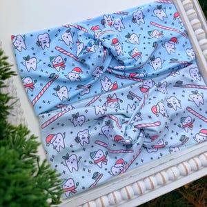 May include: Two light blue headbands with a repeating pattern of cartoon teeth wearing Santa hats and scarves, candy canes, and toothbrushes. The headbands are twisted at the top and are on a white tray. The text "LEANNALIE" is in the bottom left corner.