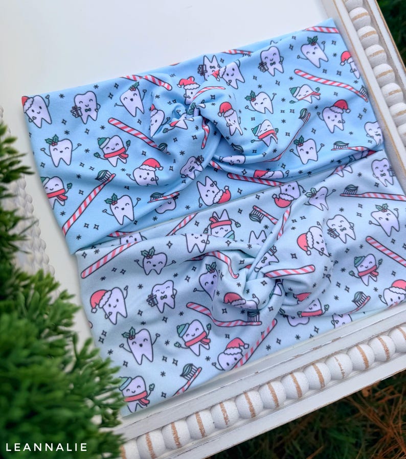 May include: Two light blue headbands with a repeating pattern of cartoon teeth wearing Santa hats and scarves, candy canes, and toothbrushes. The headbands are twisted at the top and are on a white tray. The text "LEANNALIE" is in the bottom left corner.