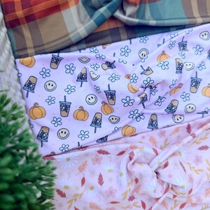 May include: Three fabric headbands with twisted knots. The top headband features an orange, teal, and brown plaid pattern. The middle headband is light pink with a repeating print of pumpkins, iced coffee cups, and smiley faces. The bottom headband is light pink with a pattern of white daisies and autumn leaves.
