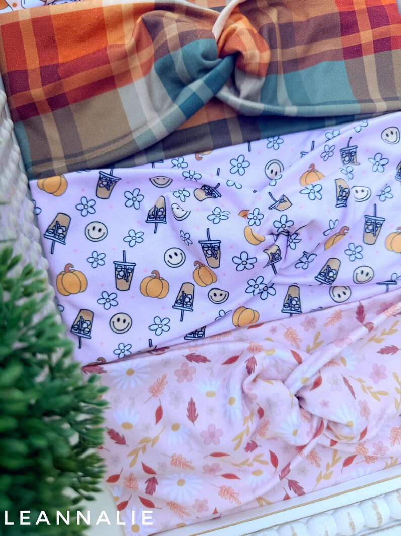 May include: Three fabric headbands with twisted knots. The top headband features an orange, teal, and brown plaid pattern. The middle headband is light pink with a repeating print of pumpkins, iced coffee cups, and smiley faces. The bottom headband is light pink with a pattern of white daisies and autumn leaves.