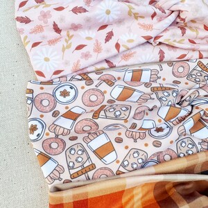 May include: Three fabric headbands with different patterns. The top headband has a pink background with white flowers and brown leaves. The middle headband has a beige background with brown coffee cups, doughnuts, and maple leaves. The bottom headband has a plaid pattern with orange, brown, and green squares.