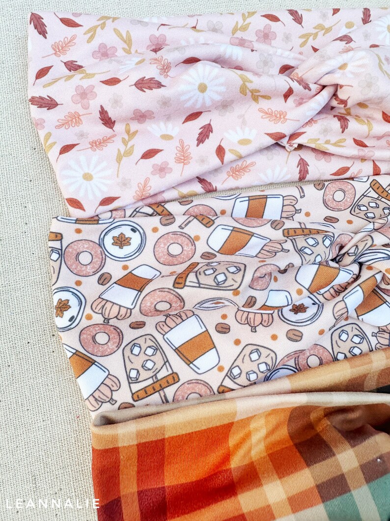 May include: Three fabric headbands with different patterns. The top headband has a pink background with white flowers and brown leaves. The middle headband has a beige background with brown coffee cups, doughnuts, and maple leaves. The bottom headband has a plaid pattern with orange, brown, and green squares.