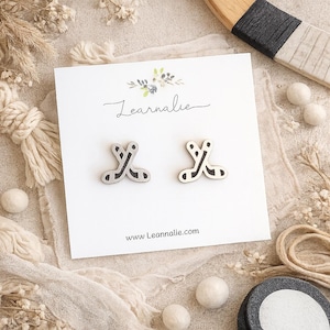 May include: A pair of stud earrings shaped like crossed hockey sticks on a white display card. The earrings are silver-toned with black detailing. The card features the text "Learnalie" in script and the website "www.Leannalie.com" at the bottom.