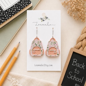 Puede incluir: Un par de pendientes colgantes con forma de mochila, con un estampado floral rosa claro en la parte delantera y un acabado de madera natural en los laterales. Los pendientes están sujetos a una tarjeta de presentación blanca con el texto Leannalie.