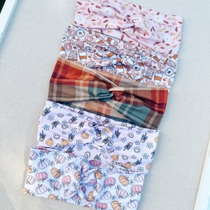 May include: Four different patterned headbands stacked on top of each other. The top headband is pink with a floral pattern. The second headband is brown with a pattern of coffee cups, doughnuts, and plaid. The third headband is a light brown plaid. The bottom headband is white with a pattern of pumpkins and flowers.