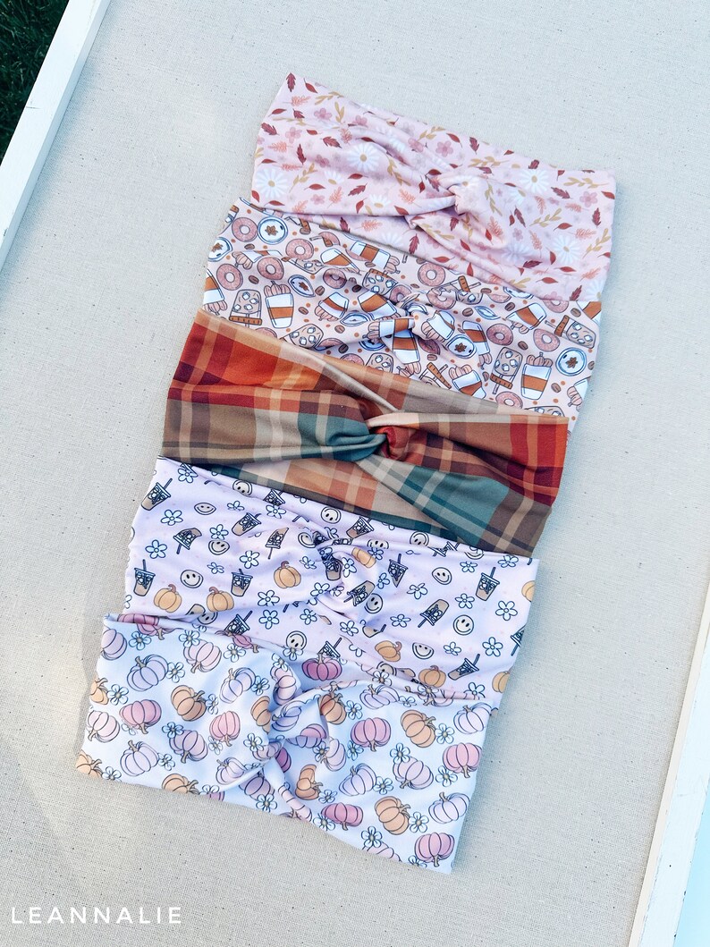 May include: Four different patterned headbands stacked on top of each other. The top headband is pink with a floral pattern. The second headband is brown with a pattern of coffee cups, doughnuts, and plaid. The third headband is a light brown plaid. The bottom headband is white with a pattern of pumpkins and flowers.