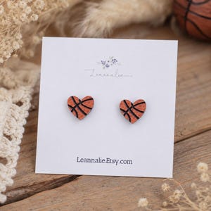 May include: A pair of stud earrings shaped like hearts with an orange and black basketball texture pattern. The earrings are attached to a white display card featuring the text Leannalie and Leannalie.Etsy.com printed in black ink.