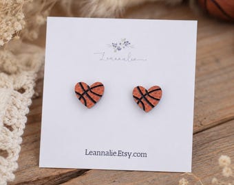 May include: A pair of stud earrings shaped like hearts with an orange and black basketball texture pattern. The earrings are attached to a white display card featuring the text Leannalie and Leannalie.Etsy.com printed in black ink.