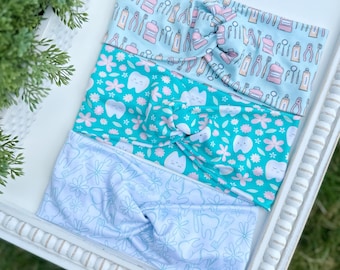May include: Three fabric headbands with a twist knot design. One is light blue with dental tools, another is teal with smiling teeth and flowers, and the third is white with teeth and flowers.