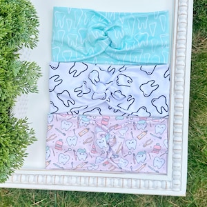 May include: Three fabric headbands with dental-themed patterns. The top headband is light blue with white teeth, the middle is white with black teeth, and the bottom is pink with dental tools and smiling teeth.