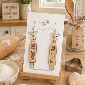 May include: Wooden rolling pin earrings with silver bow accents. The earrings are shaped like miniature rolling pins and have the words "MADE WITH LOVE" engraved on them. The earrings are displayed on a white card with the text "Leannalie.Etsy.com".